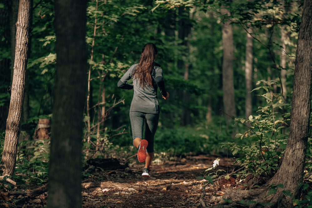 Exercising outdoors is healthy for active lifestyle runners. Autumn trail run woman running in nature from behind in dark forest.