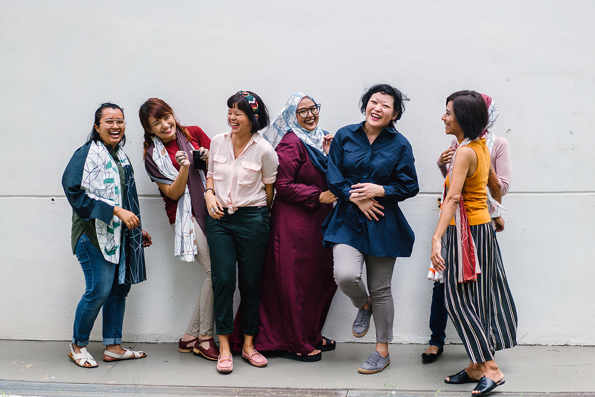 A diverse group of Asian women (Malay and Chinese) goof around with one another against a white wall. They are leaning on one another in a row and are smiling and laughing at one another.